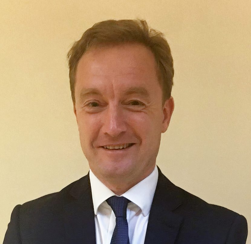 Headshot of David Alexander in a black suit jacket, white shirt and navy blue tie