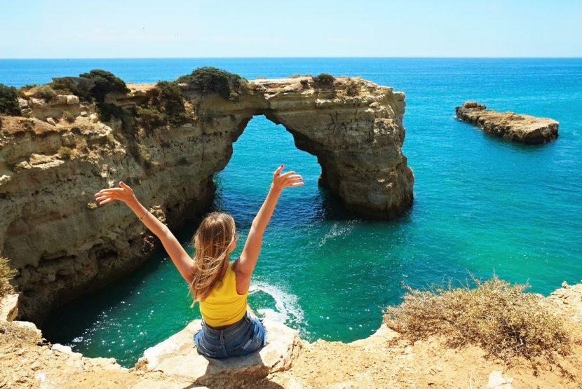 Woman with long blonde hair and yellow vest top sat on rock facing blue, green ocean with arms raised in the air