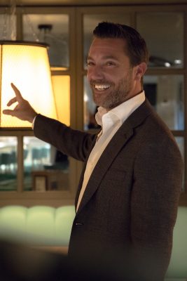 Chef Gino D'Acampo stands in a dark suit jacket and white shirt pointing behind him, smiling off camera