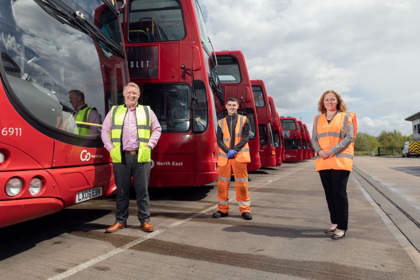 To left of image, red double-decker busses in a line. In foreground three people equally spaced out. On the left is Colin Barnes in a yellow high-vis waistcoat, in the middle is apprentice Oliver Barry in full body orange high-vis and on the right is Suzanne Slater in an orange high-vis vest