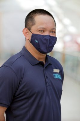 Man with short dark hair, wearing a mask and dark blue polo shirt with Metrocentre logo and name badge on his left breast