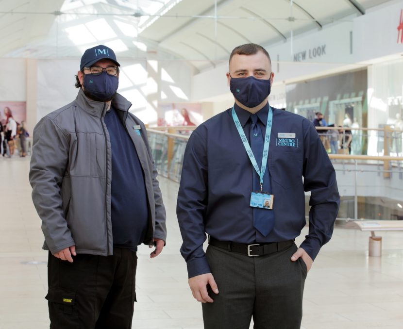 Two Metrocentre staff, both wearing masks. One is wearing a dark blue shirt with a lanyard around their neck with one hand in their suit trouser pocket. The other is wearing a cap and grey jacket