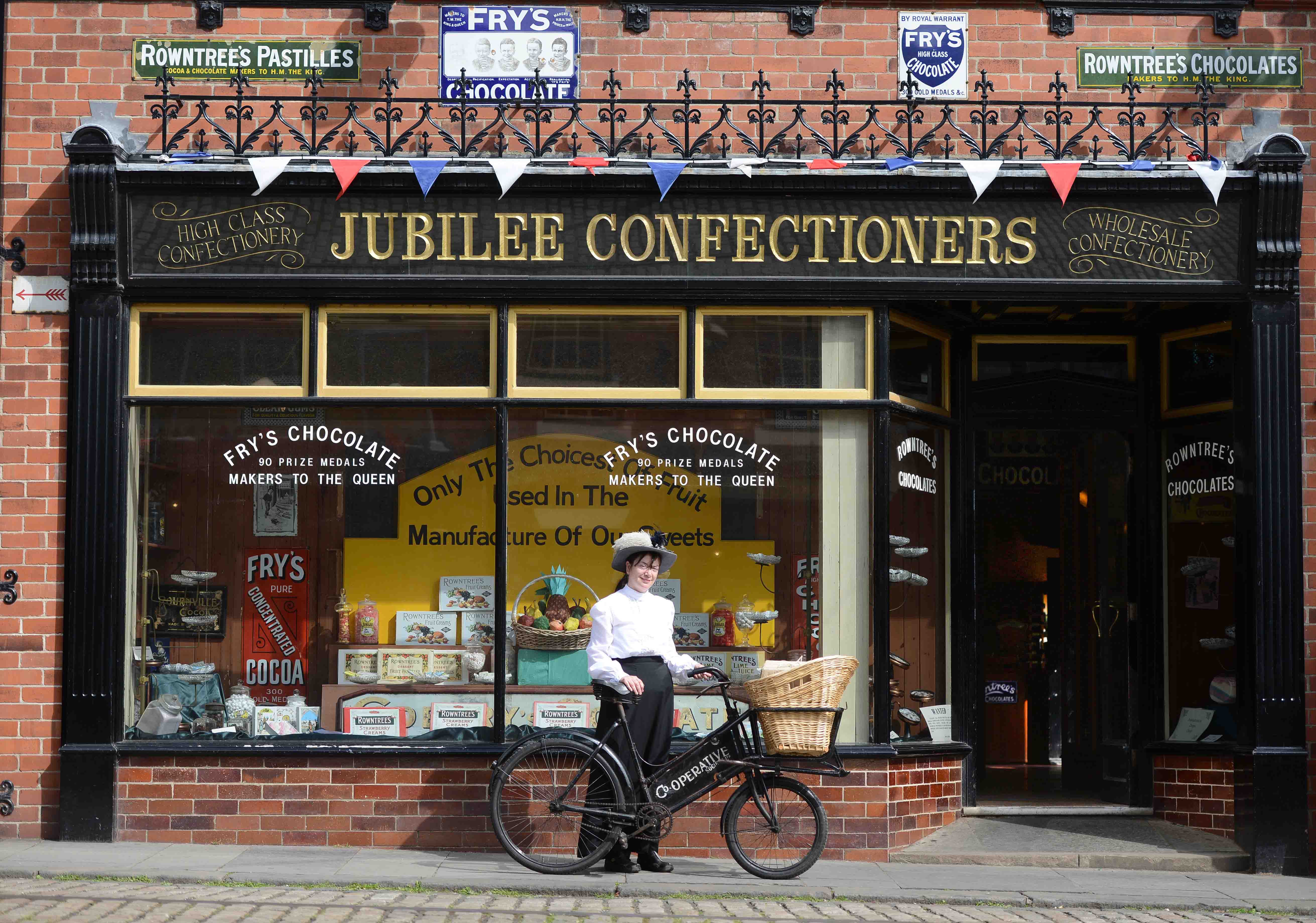 Exterior of Jubilee Confectioners. 'Jubilee Confectioners' is written in gold on a black background above the shop window. Woman in traditional Edwardian dress holding a vintage bicycle stands in front of window.