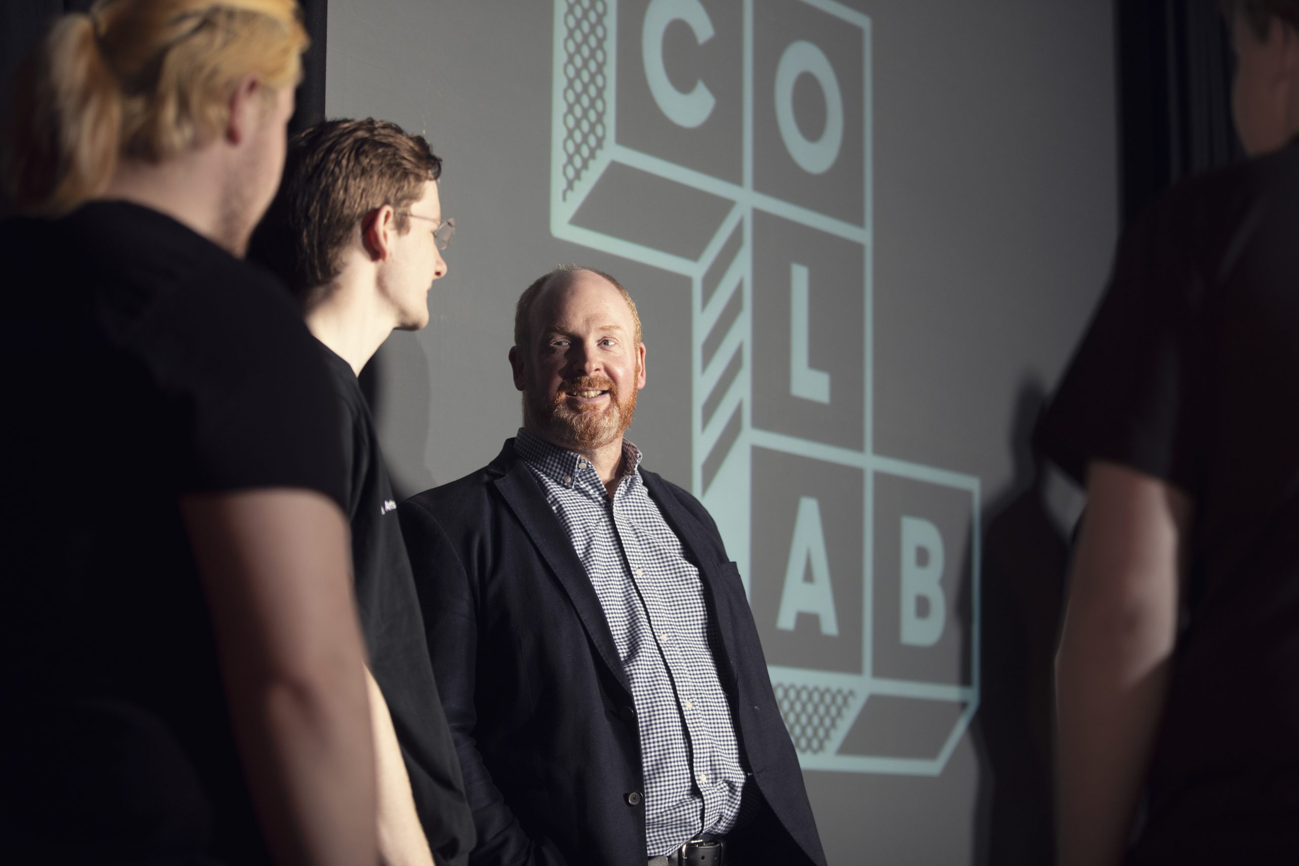 Man with beard in checked shirt and blazer in front of screen with CoLab written within geometric shape. Man is surrounded by other people