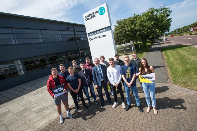 Group of young people stood in semi circle in front of Gateshead College sign with building on the left and trees on the right in the background