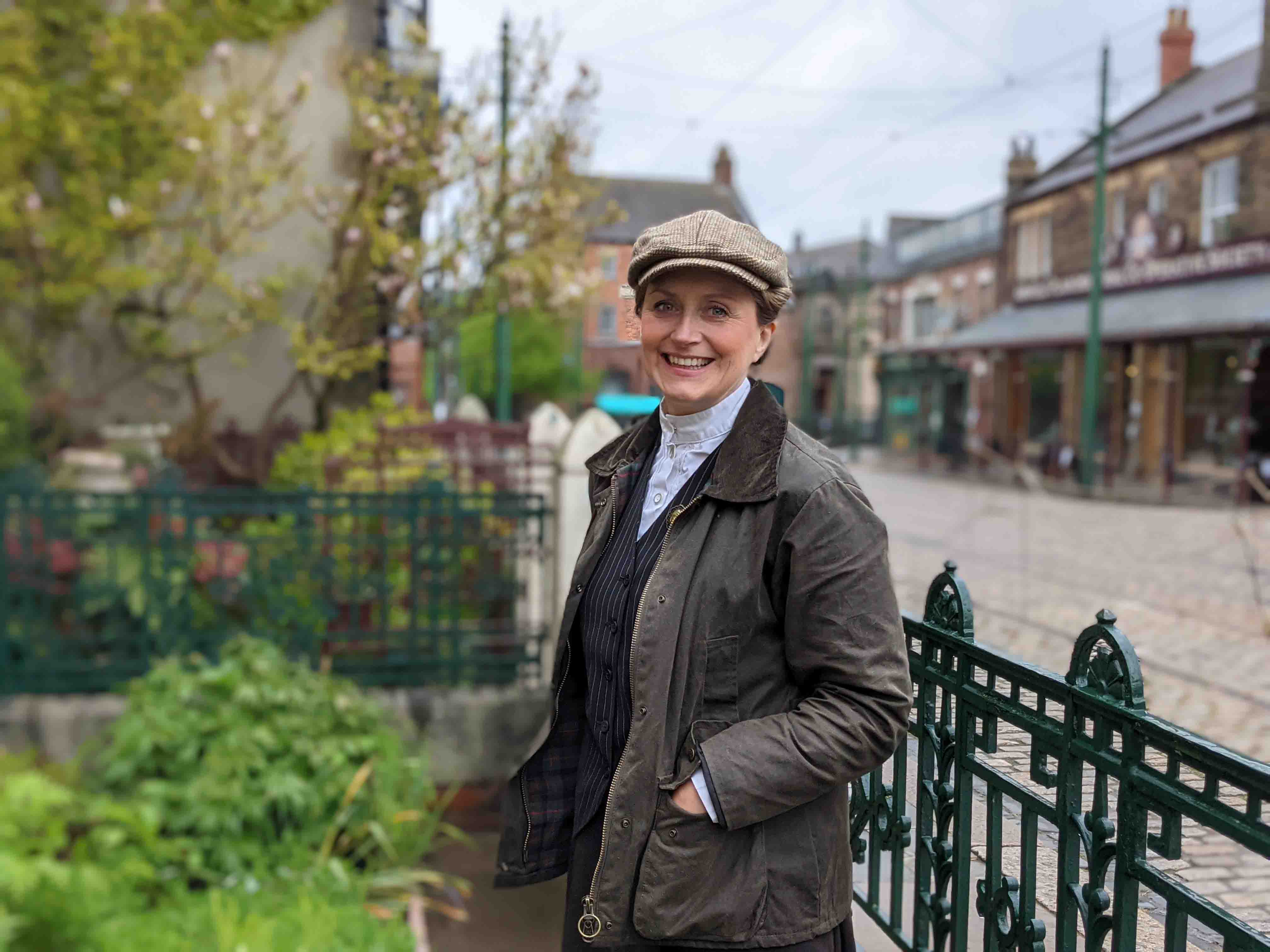 Woman leaning against black railings dressed in dark jacket, shirt and flat cap