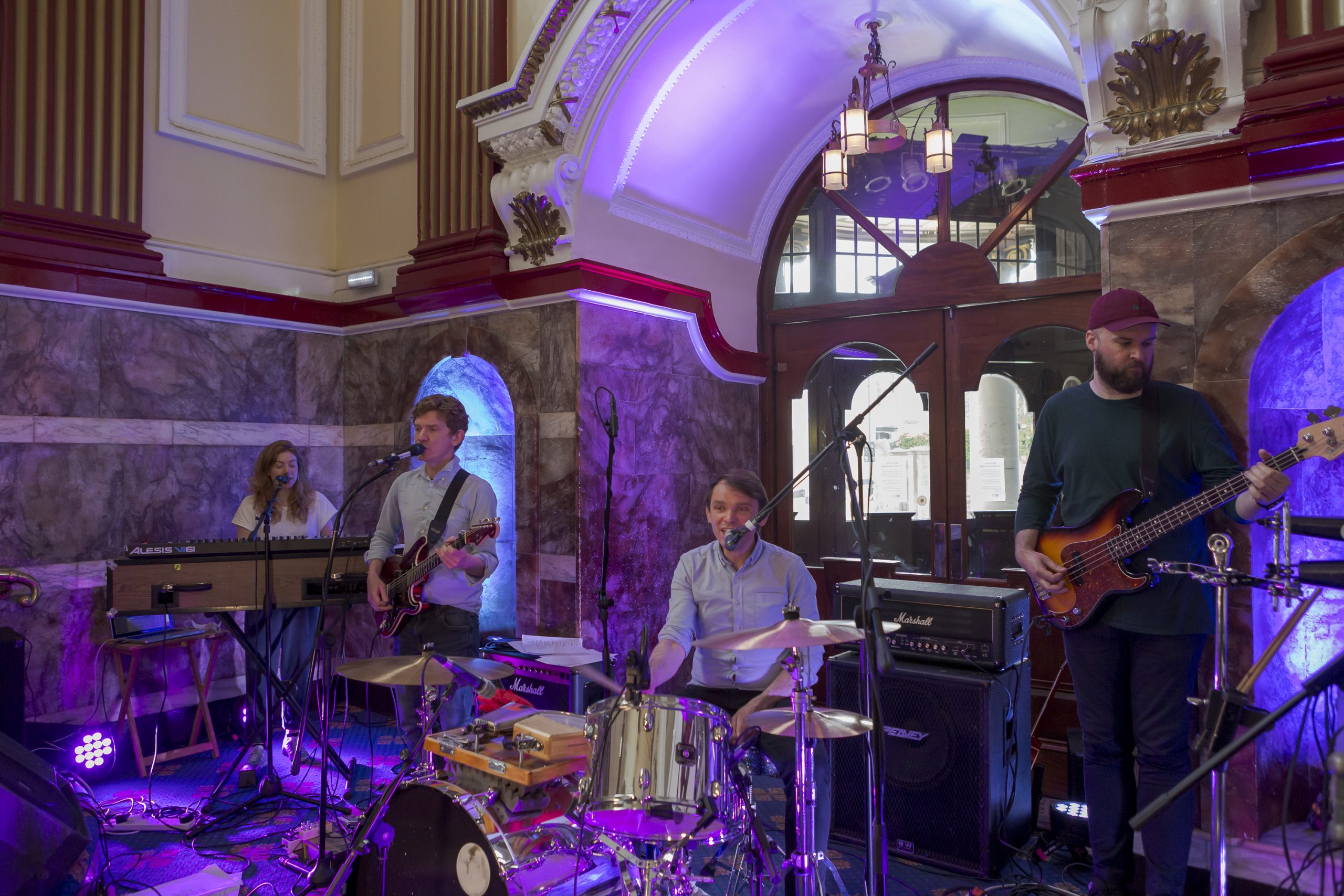 Band with guitarists, drummer and keyboard player performing. Keyboardist on the far left is singing and has long brown hair, guitarist is stood next to her in blue shirt and is also singing. Drummer sits in the middle with drum kit in a blue shirt and short brown hair singing. Bass guitarist stands on the right wearing red cap and has a dark beard.