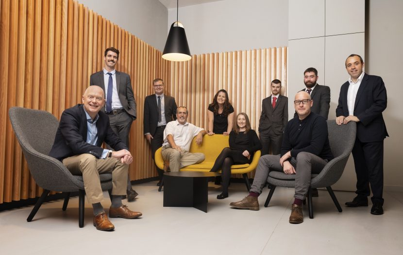 Group of 10 people sit and stand in corner of a room. Wood panelling lines the bottom half of the walls. From left to right: a man with no hair sits smiling at camera wearing a navy blue blazer and beige trousers, next to him stands a man with dark hair and moustache, in a grey suit with light blue shirt and tie. Stood in the corner is a man with light hair and glasses wearing a dark suit and tie with a light shirt. Next to him sits a man with white hair and facial hair with glasses in a white shirt and beige trousers. A woman stands leaning behind the yellow sofa with dark hair and black short-sleeved dress. Next to her sits a woman with light hair and black dress and tights. Behind her stands a man with dark hair and facial hair, with his hands in his grey suit pockets. He is wearing a white shirt and red tie. Next to him stands a man with dark hair and beard with a dark suit. The next man sits in front wearing glasses, a black top and grey trousers, he has little hair. The final man stands behind with dark hair, a dark suit and white shirt.