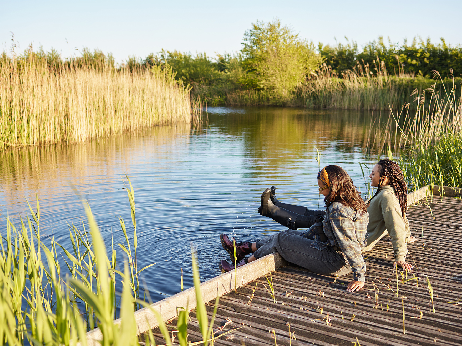 Two women sat on a jetty surrounded by water and grass reeds