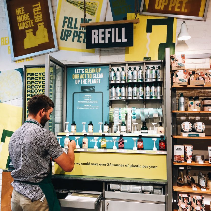 Product refill station in Body Shop store. Man to the left is filling up a bottle at the station. Surrounded by bold and eyecatching green and blue signage. Lines of aluminium bottles are at the top of the station.