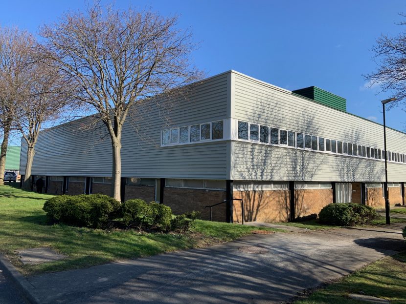 Large, wide, low, boxy grey building set atop brick foundations/floor level. Grass, bushes and a few trees are dotted around outside in the foreground.