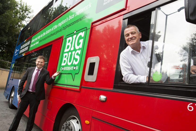 Man dressed in dark suit and purple tie stands beside red double decker bus gesturing towards advert on bus for the Big Bus Conversation. Another man sits in the drivers seat of the bus in a white shirt. Both are smiling towards the camera.