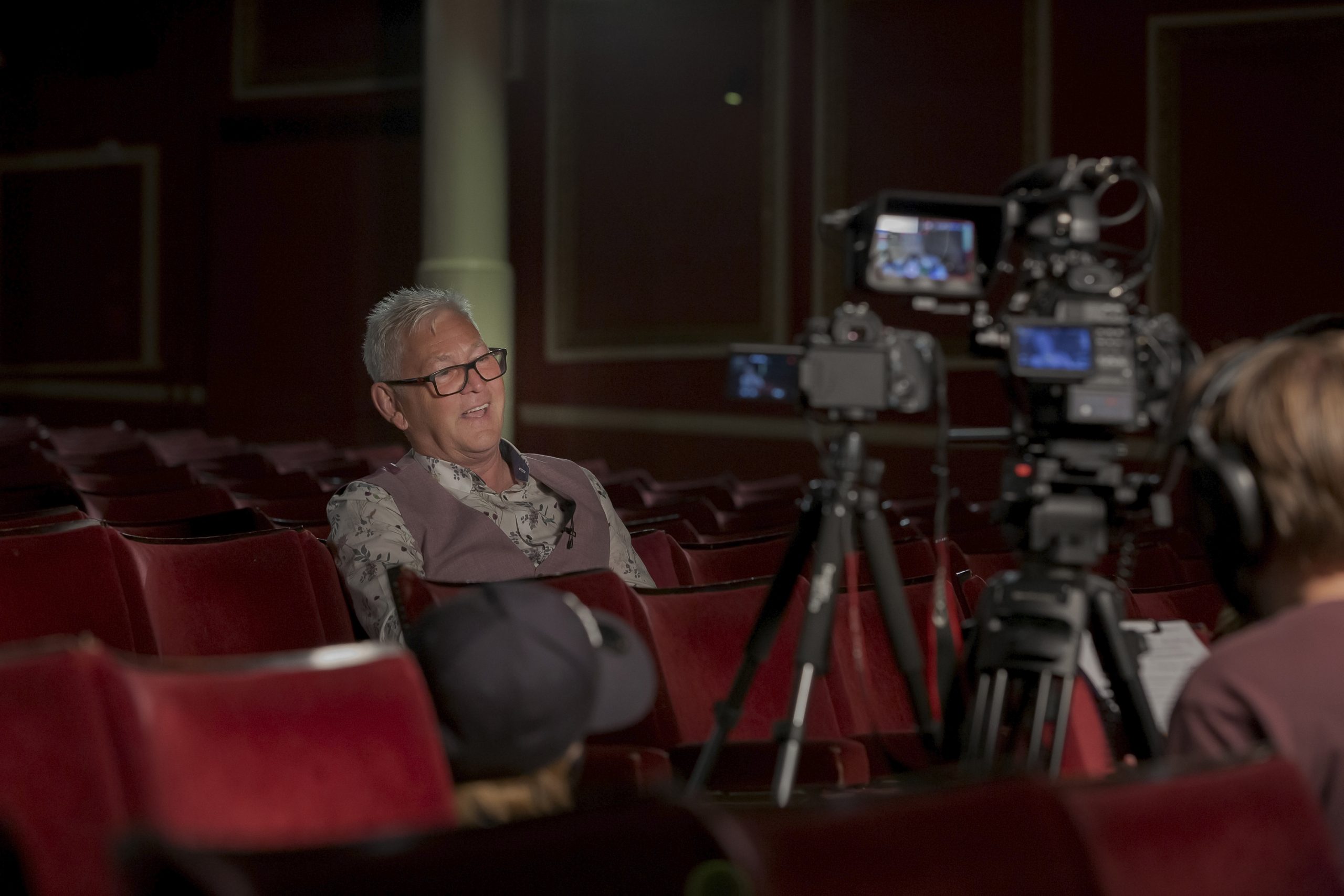 Steve Colman from Smooth Radio pictured during the Your Theatre Your Story event at the Empire Theatre. He sits in a red theatre chair in a purple waistcoat with dark rimmed glasses on and short white hair. A large filming camera with lots of wires and a camera operator with headphones on sits in the foreground on the right hand side of the screen