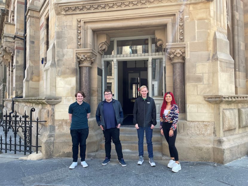 Four young people stand outside a sandstone building. On the far left stands a man with short brown hair wearing a navy short-sleeve top and dark trousers, next to him stands another young man with short dark hair and glasses wearing a blue jacket and dark trousers, next to him stands another young man slightly taller than the rest with longer blonde hair, a dark jacket and blue jeans, next to him stands a young woman with bright red hair a patterned jacket and dark skinny jeans and white chunky trainers