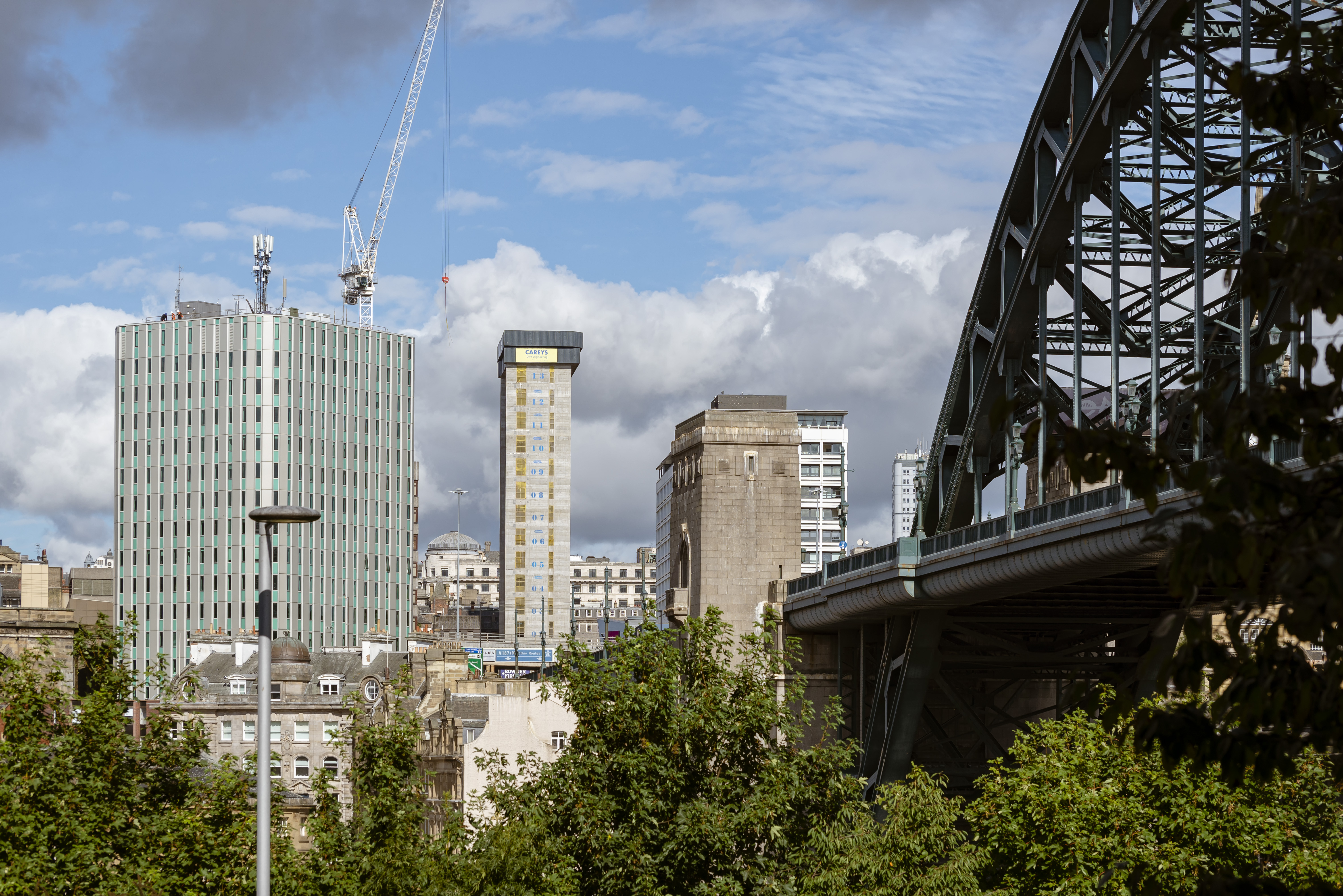 Skyline image of Newcastle city centre. The green Tyne bridge is nestled to the right of the image and the new grey concrete of the Bank House building stands tall in the middle of the image with a large crane towering over it to its left. There are green tree tops in the foreground of the image.