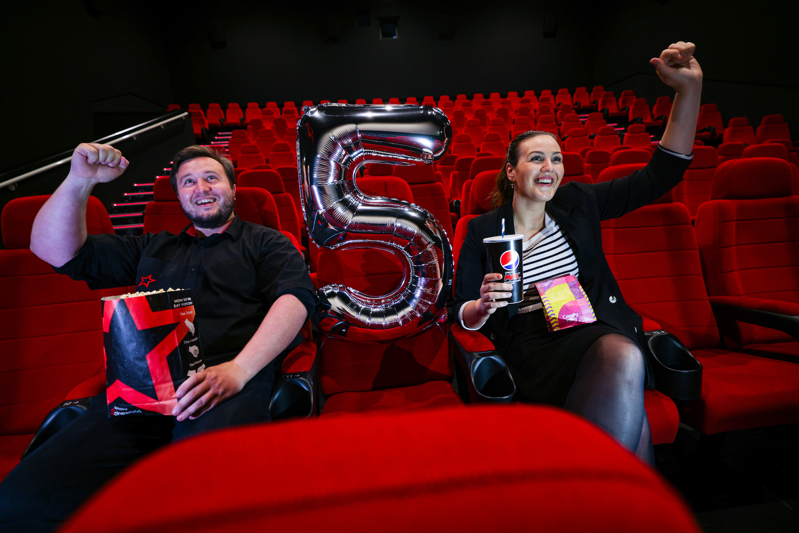Man and woman sit in a movie theatre on red velvet seats. On the left sits the man with short brown hair and beard in all black clothing. He holds a bag of popcorn and has one fist up in the air in a joyful pose. On the right sits a woman with dark hair slicked back in a ponytail wearing a striped top and black blazer holding a drink in one hand and with fist up in the air like the man. Both are smiling and there is a '5' balloon placed in between them.
