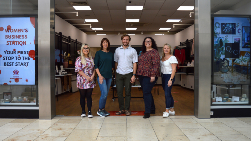 5 people stand in the doorway of a glass fronted store smiling towards camera. On the far left is a woman with long blonde hair and dark glasses, wearing a short summer dress with black leggings, next to her is a taller woman with dark brown bob haircut in a long sleeve blue top and blue jeans. Next to her is a taller man with dark, short, curly hair wearing a light polo shirt, next to him is a woman with long, dark, curly hair, long sleeve floral top and dark trousers. On the right hand side is a shorter woman with blonde hair, wearing a short sleeve white blouse and blue jeans.