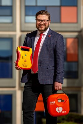 Sergio Petrucci - man in dark blue suit, white shirt and red tie stands holding two defibrillators, one in each hand