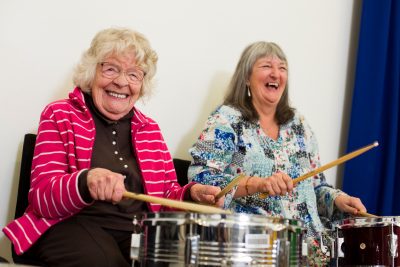 Two older women laughing and playing the drums. One the left the woman is wearing a pink fleece and black turtle neck jumper and has short white hair. On the right is a woman with longer, grey hair in a blue patterned top