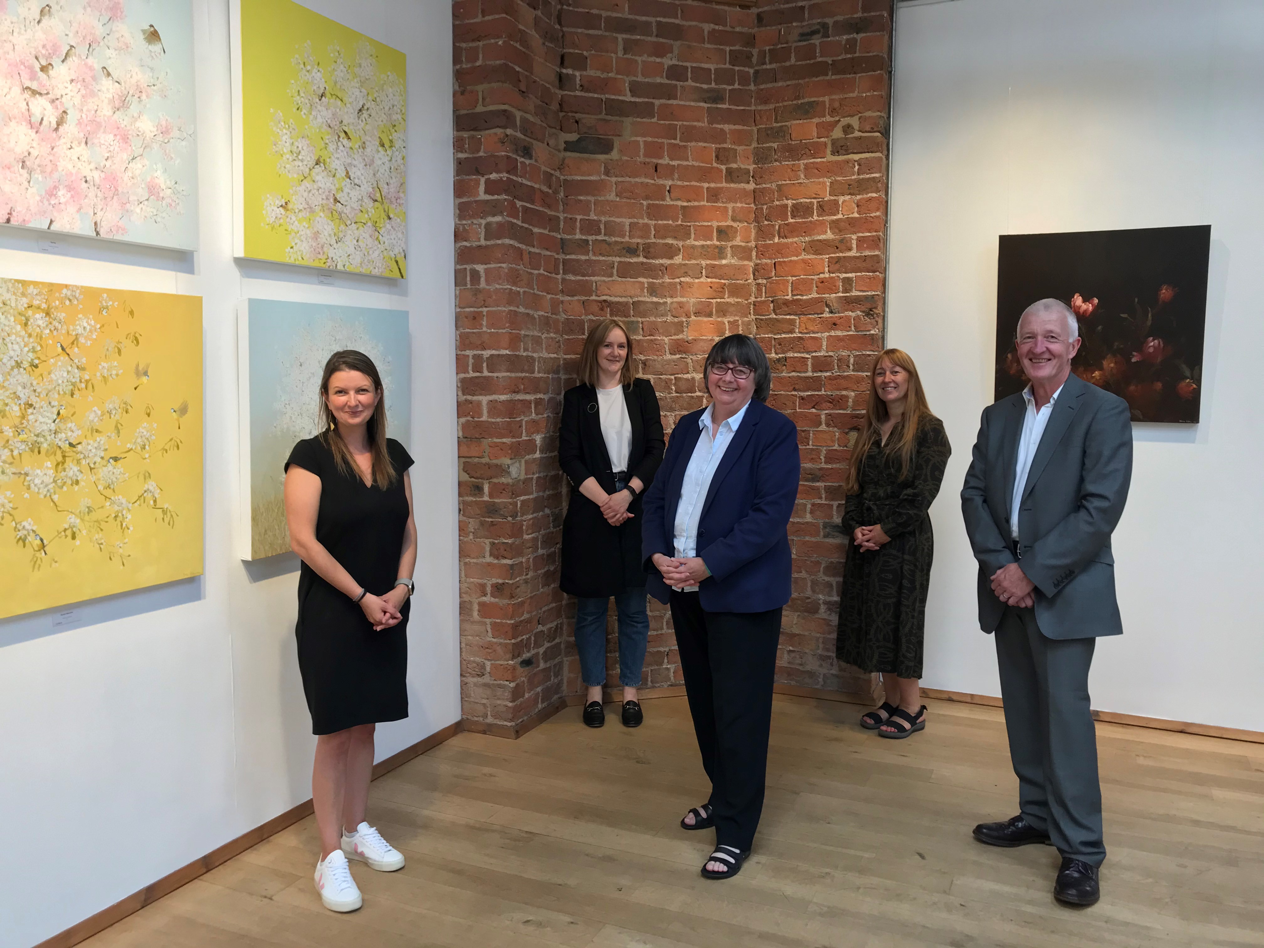 Four people stand spaced out in art gallery. On the left wall there are four large square paintings in pastel colours. Next to the wall stands a small woman in a black short sleeve dress and white trainers smiling towards camera. Next to her stands another woman in a dark blue blazer, white blouse, dark trousers with glasses also smiling to camera. On the right of her stands a man in a grey suit and white shirt. He has short white hair. Behind him stands a woman with long hair and wearing a longer dark dress and sandals. Next to her to her left stands a woman with a bob haircut wearing a long black blazer and denim jeans.