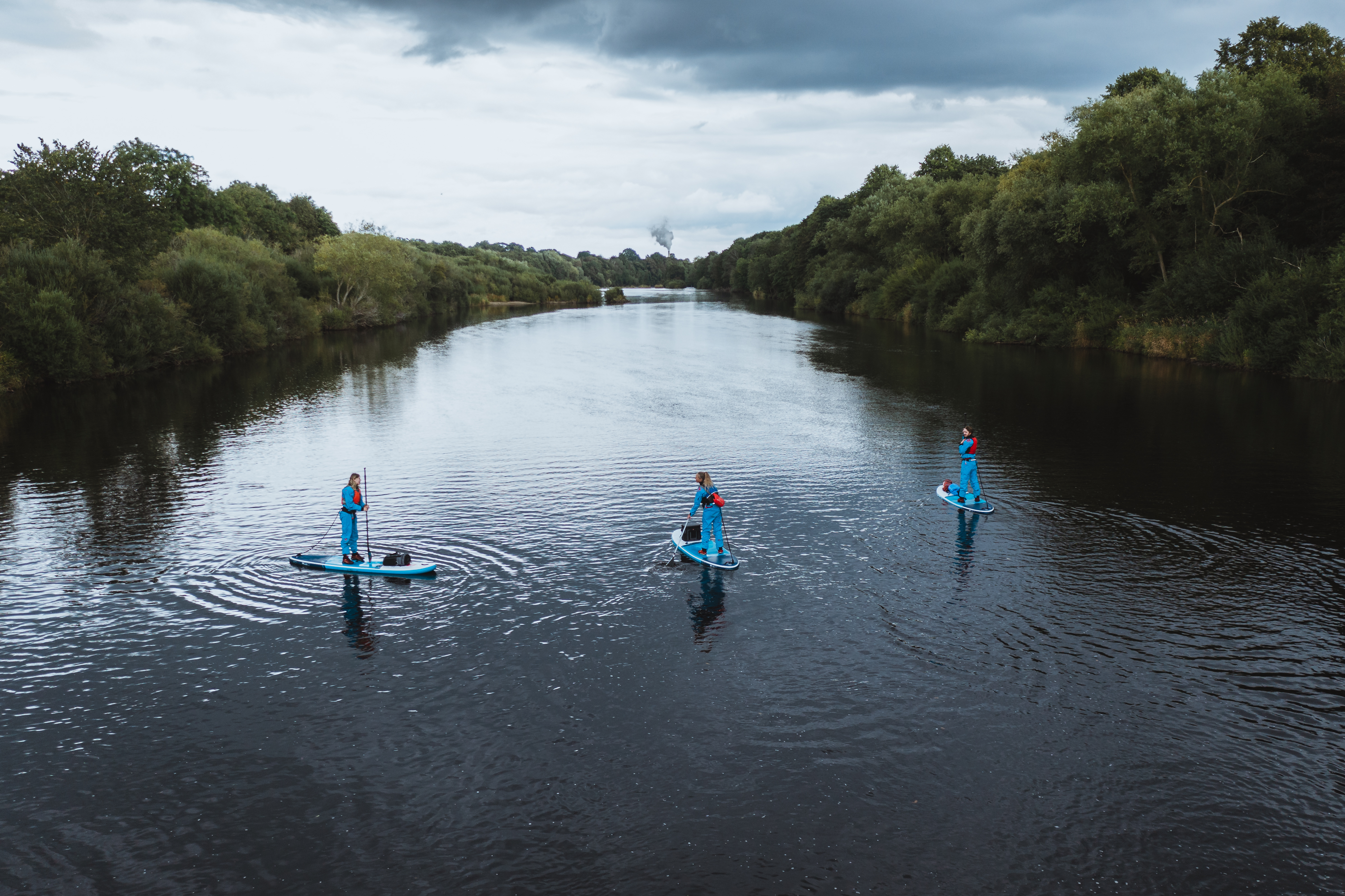 Shot of river surrounded by thick green trees, there are three people stood on top of blue paddle boards on the water