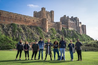 Group of people stand in horizontal line in front of a large castle that is on a cliff edge behind them. In the centre is Rob Lundgren dressed in Viking costume and with a sword in between his hands