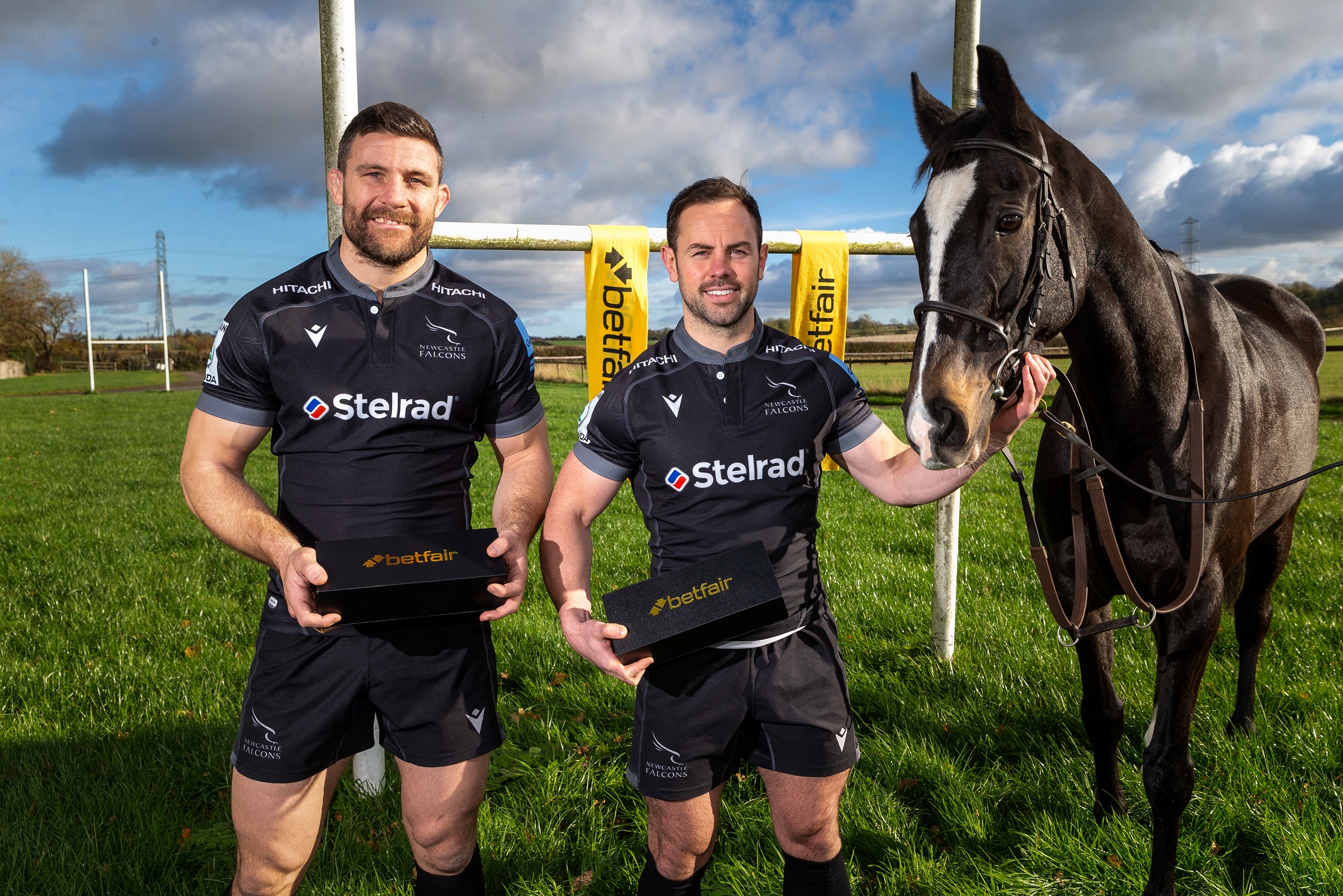 Two men and a black horse stand in front of rugby goal posts in a field of bright lush green grass. The rugby players aredressing in black and grey rugby kit and one is holding onto the horse.