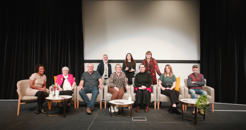 Group of people on stage. Seven people are sat on comfy chairs and in the second row three people are stood