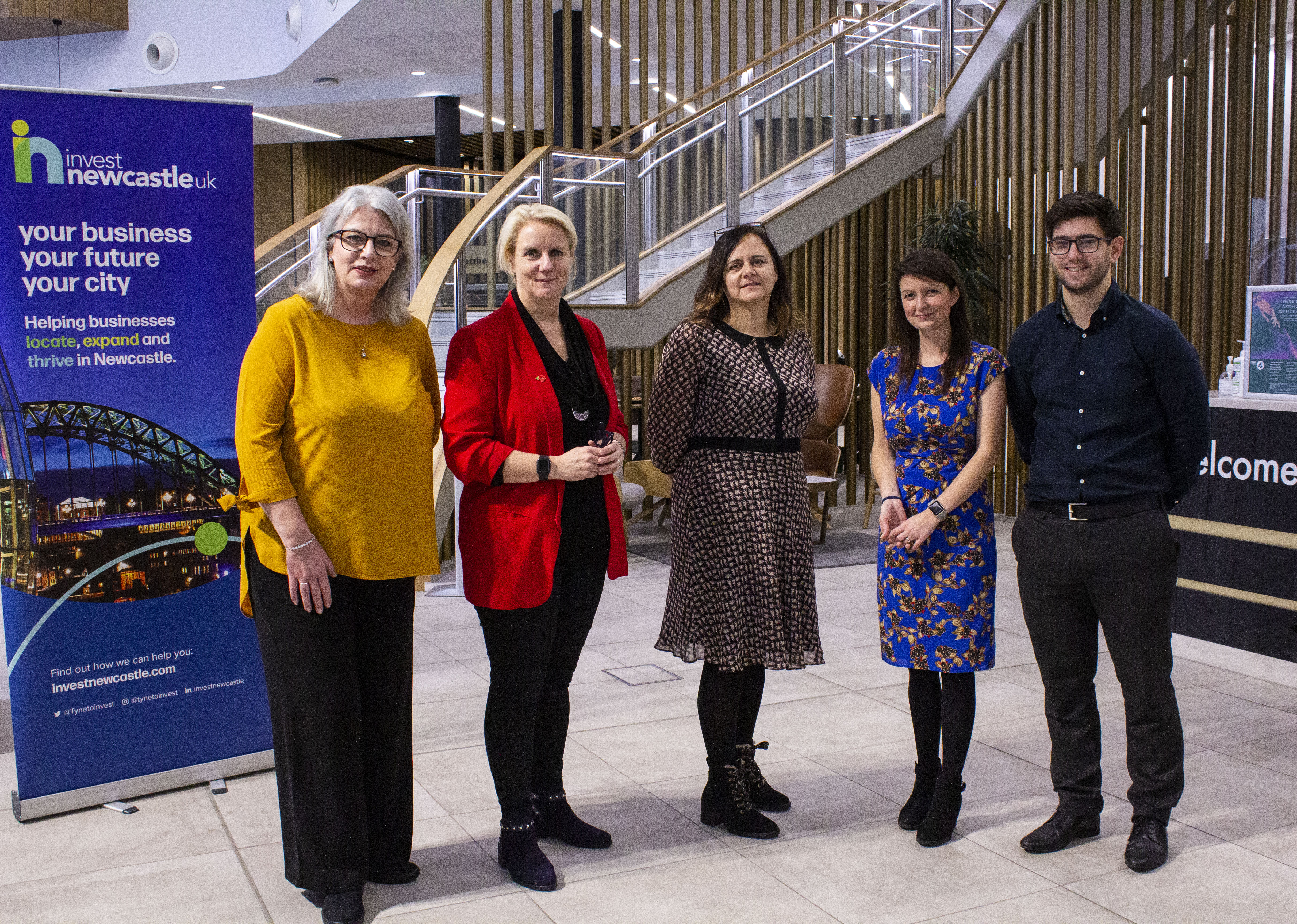 Five event organisers stand in a front of an Invest Newcastle banner and in front of wooden spiral stairs