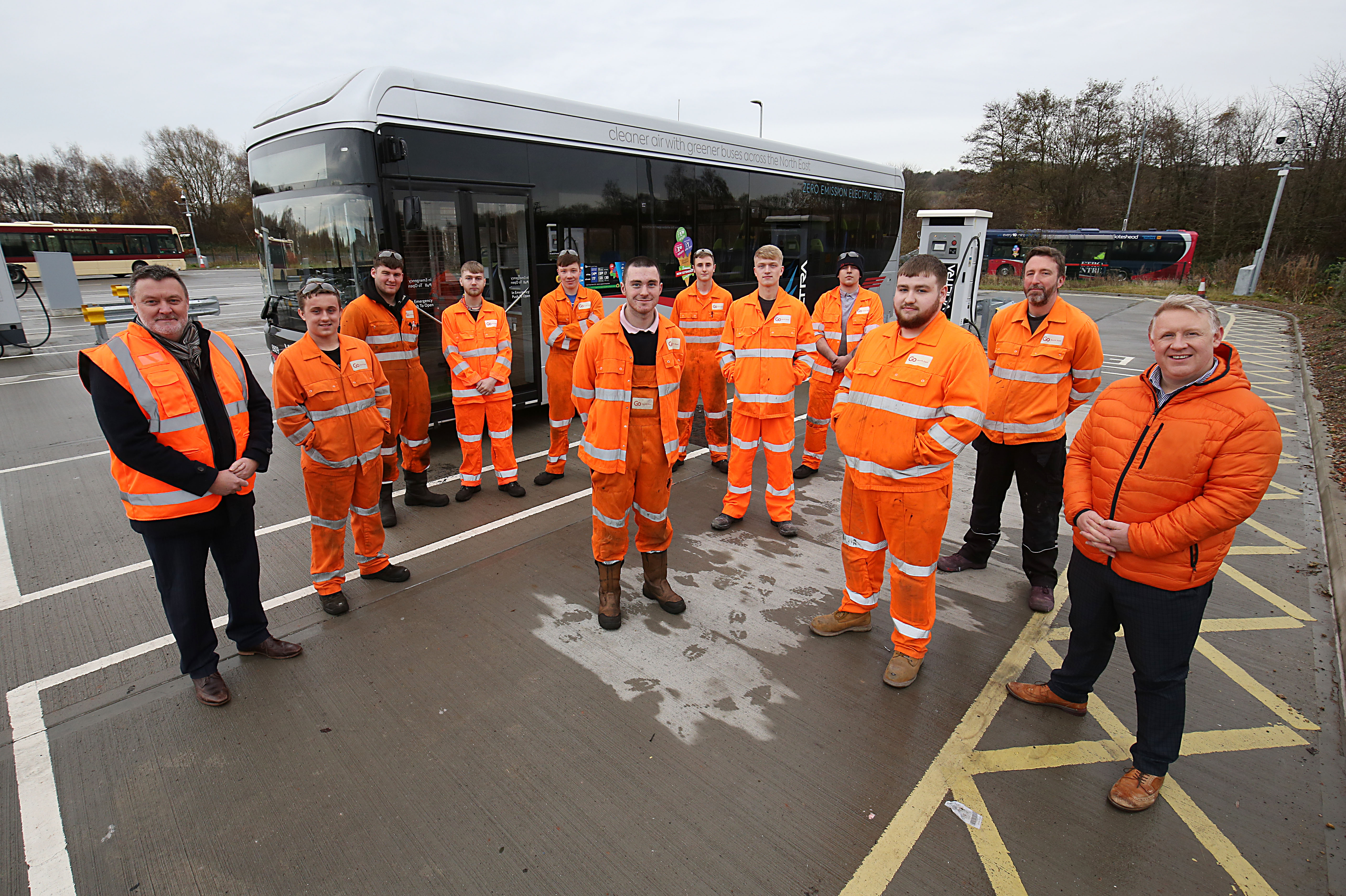 Group of 12 people stood in front of a silver bus all in orange high vis