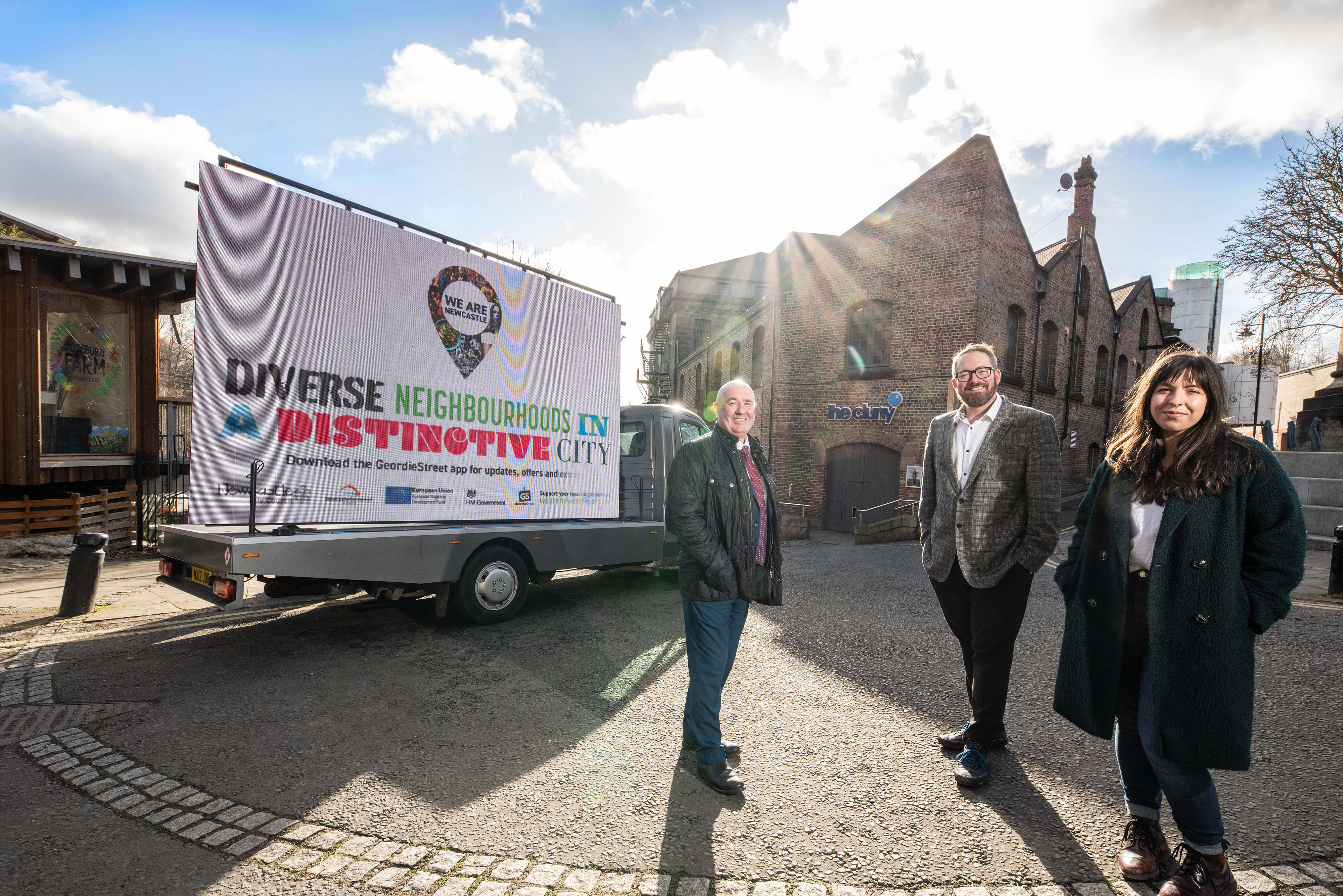 Three people stood next to a van with a large poster attached to the side promoting the Ouseburn neighbourhood