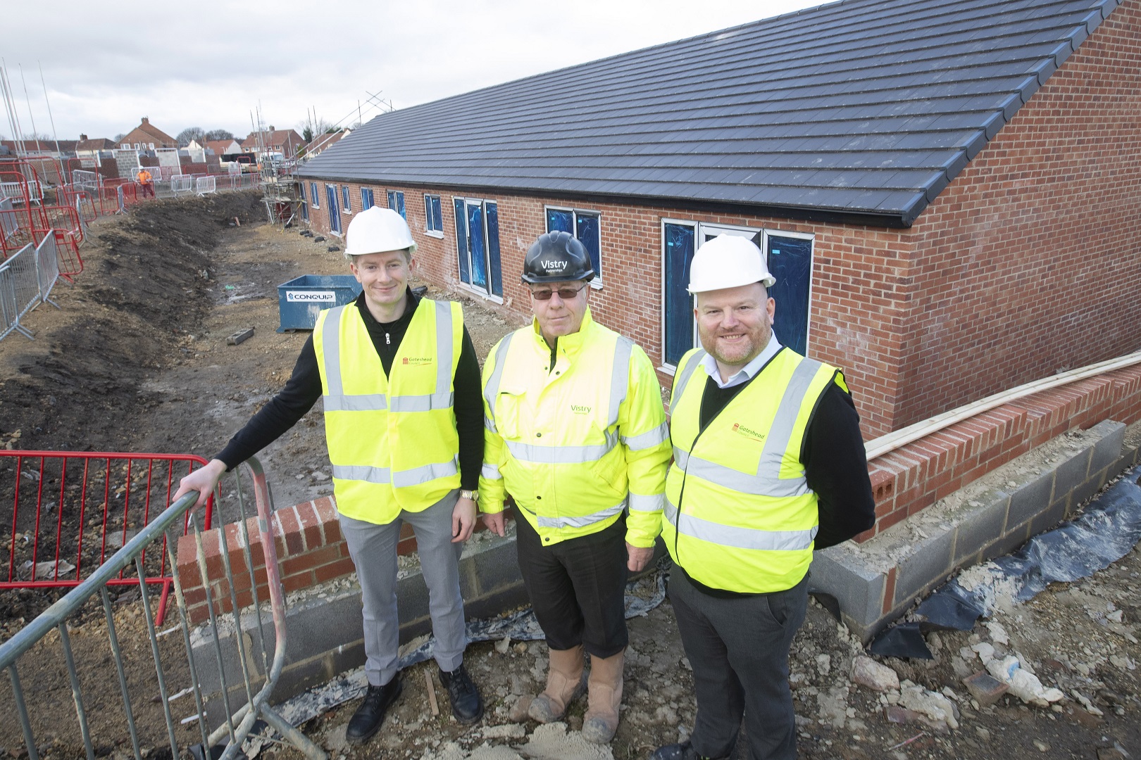 Three people in hi-vis vests and hard hats stood in front of housing development