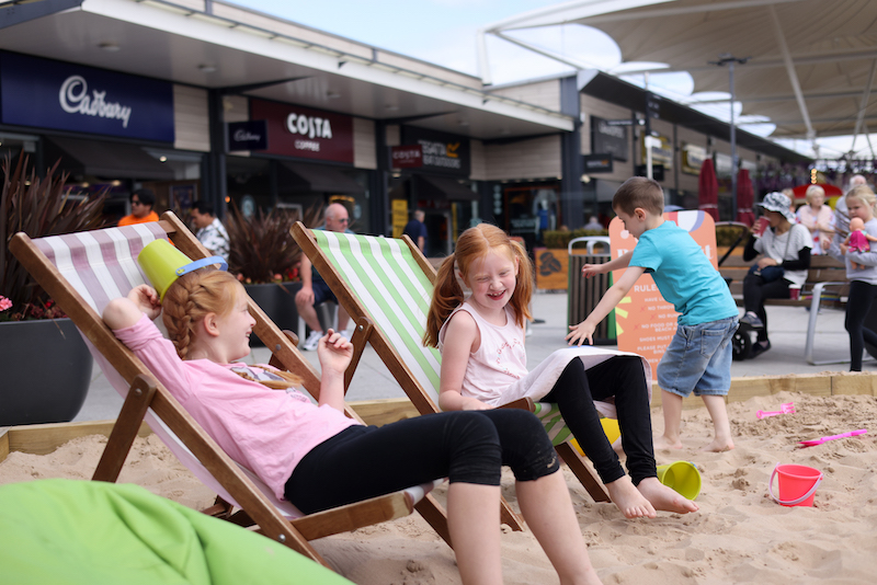 Two girls sat in deck chairs in large sandbox outside stores in Dalton Park Outlet