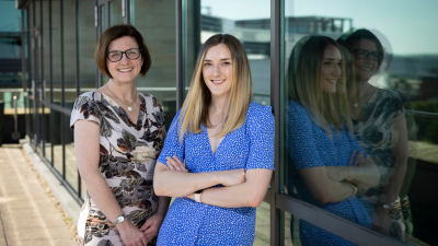 Two women stand against a window. On the left a woman with short dark hair and glasses and on the right a woman with longer and lighter hair stands with arms folded. Both smiling