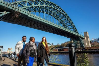 Three people walking along the river Tyne in warm jackets under the green Tyne Bridge