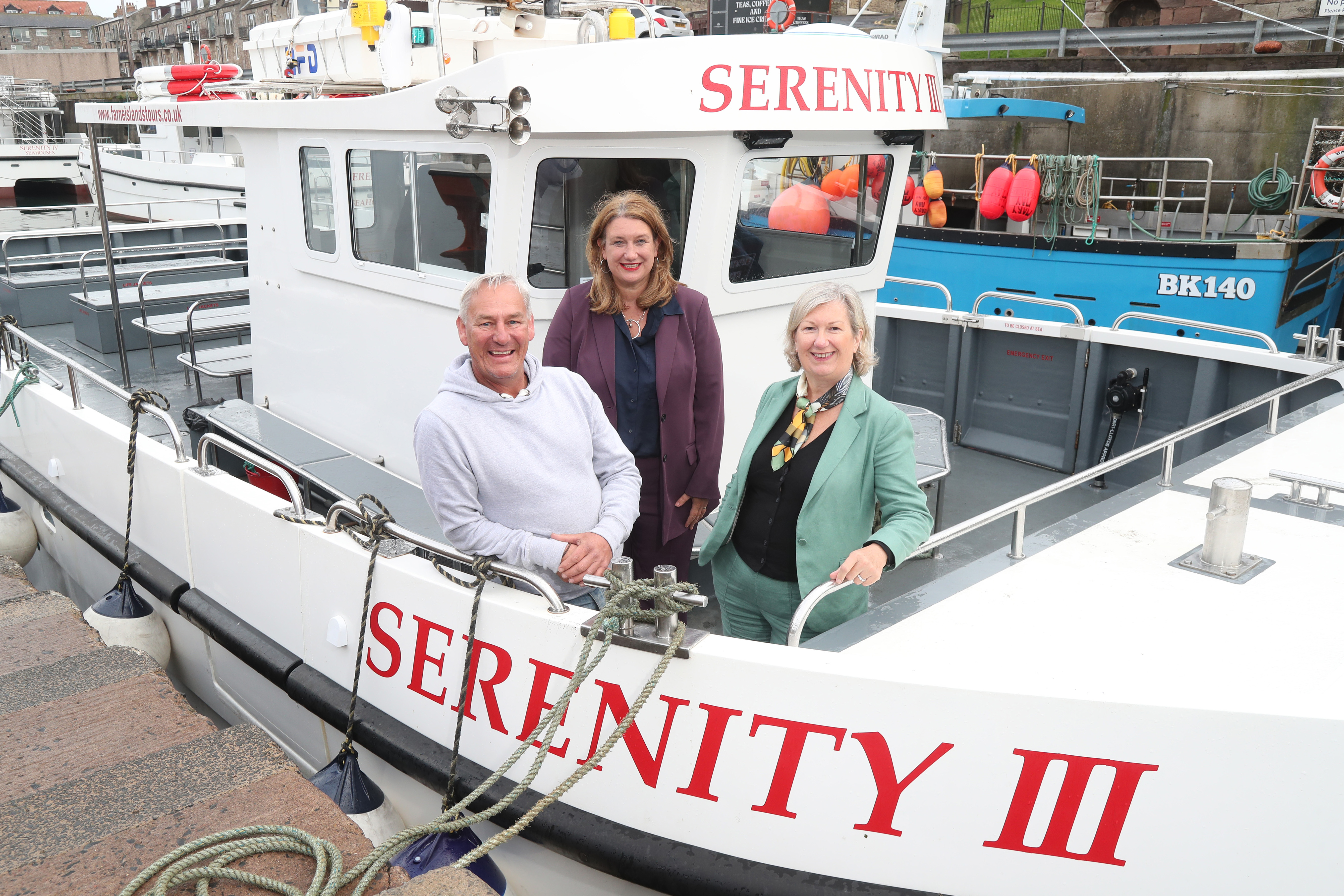 Three people smiling to camera stood on boat which is docked