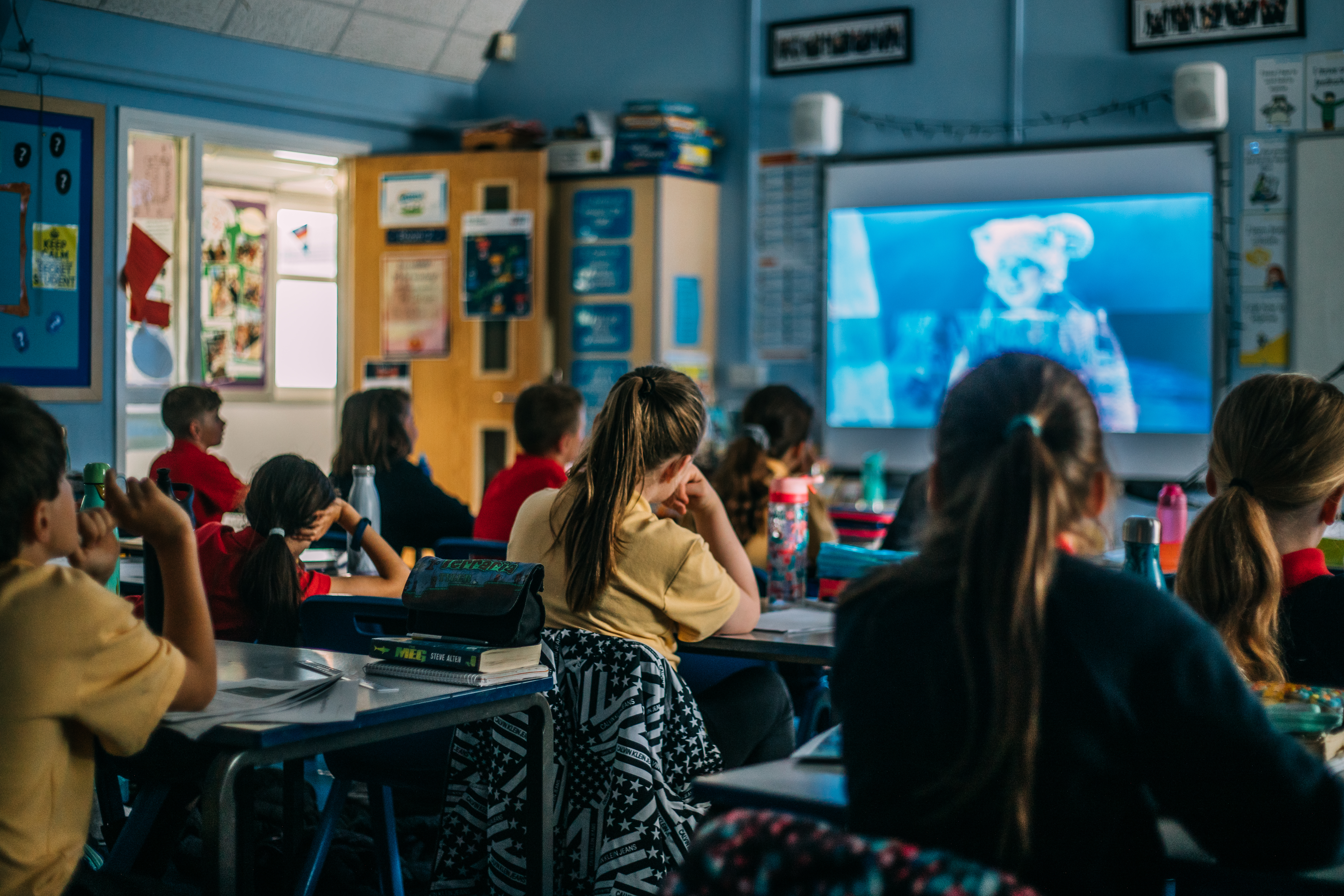 Photo from back of classroom, young school children looking towards screen at the front of the room.