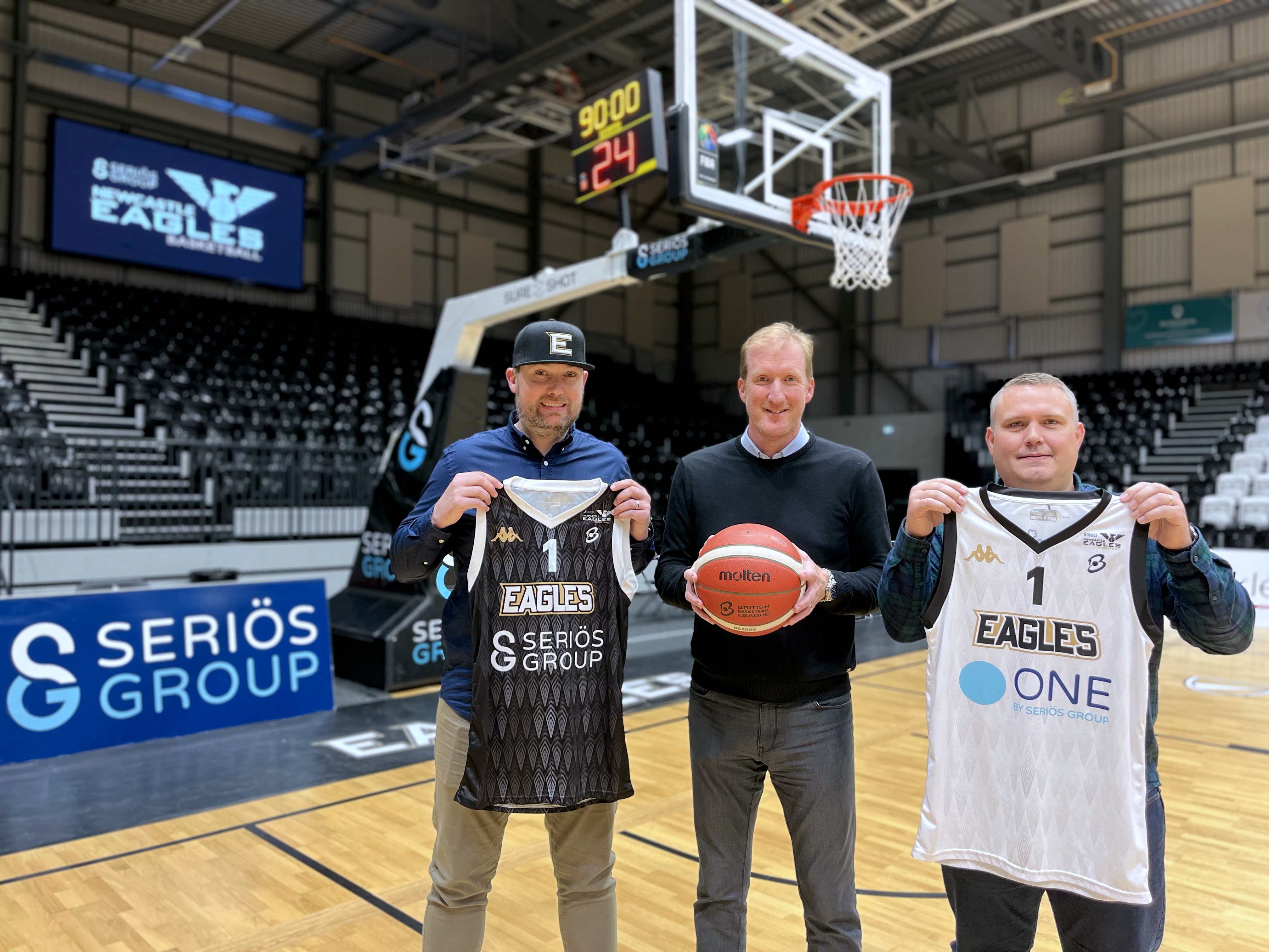 L-R Lee, Paul and David standing on the Newcastle Eagles basketball court, smiling at the camera. Lee and David are holding black and white Newcastle Eagles basketball jersey. Paul is holding a basketball.