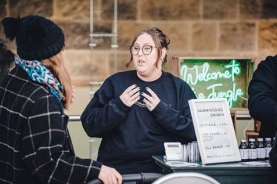 Lady wearing glasses from a company called Hummingbird Drinks talks to a customer at a market stall