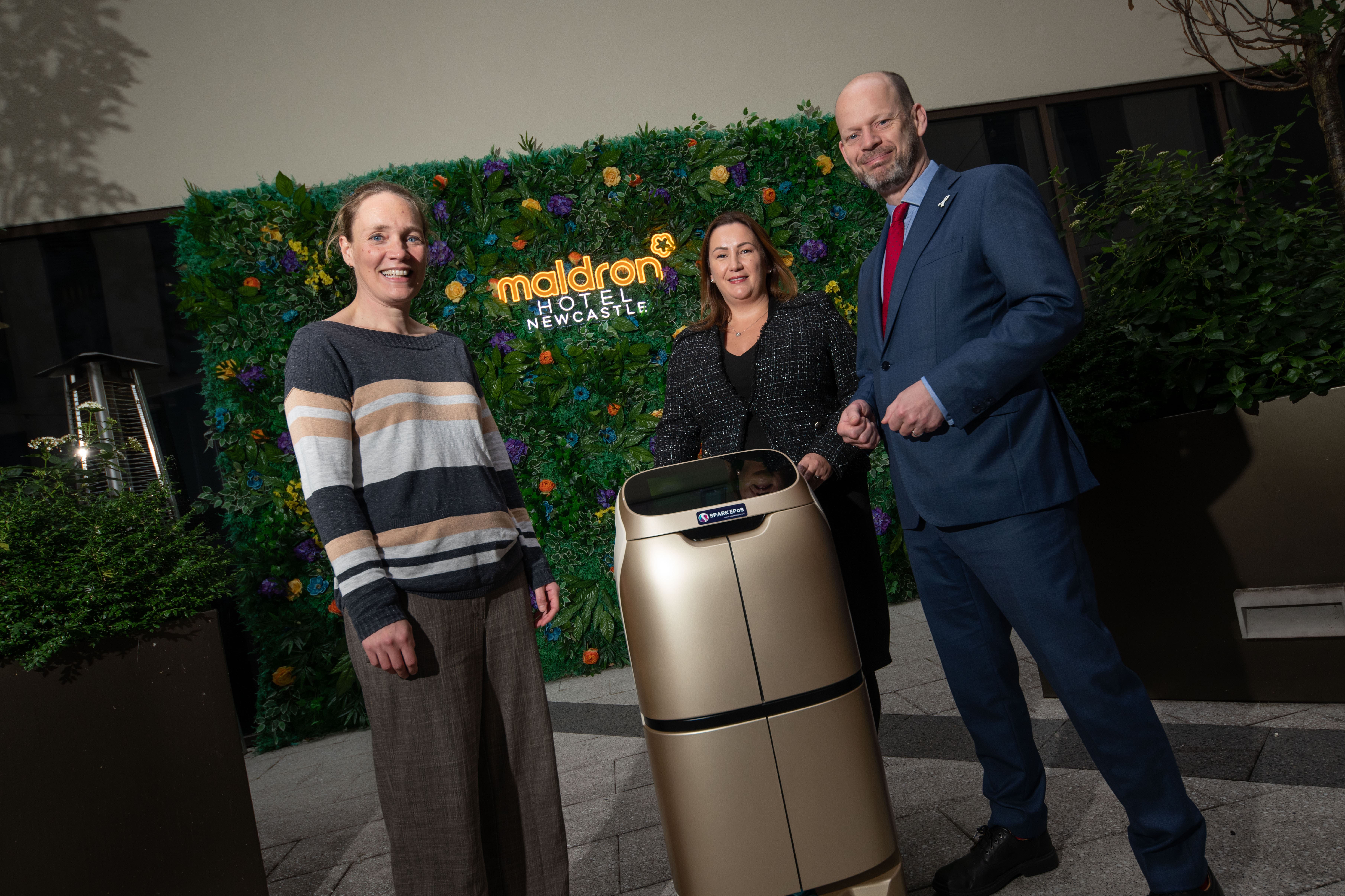 Shona, Anna and Jamie stood around the robot with background of greenery and Maldron logo