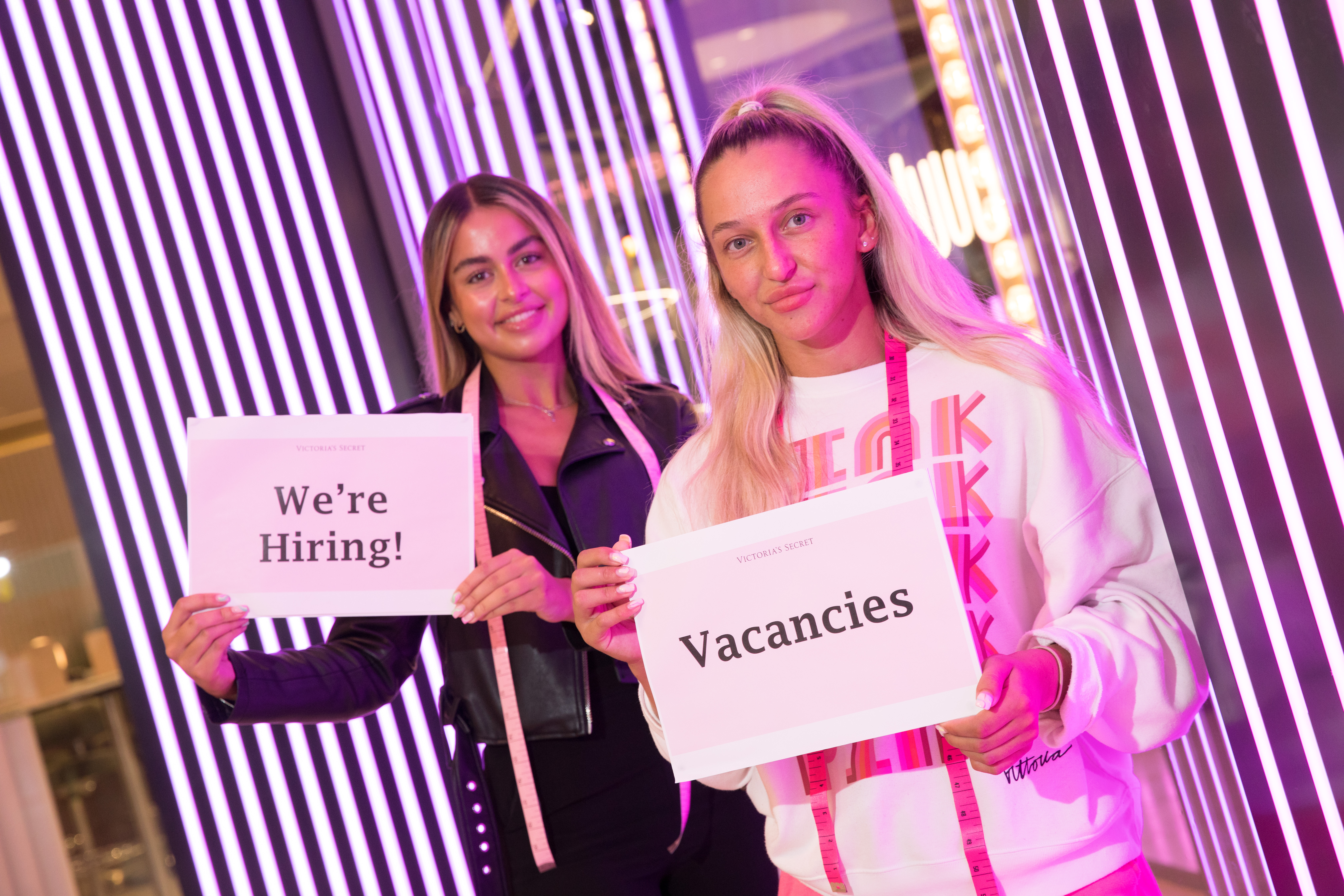 Two women stood in front of vertical, pink fluorescent lights holding signs saying: 'We're hiring!' and 'Vacancies'