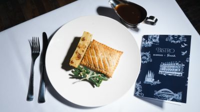 Moody image of a steak bake on a plate with potatoes and bundle of green beans with Greggs x Fenwick Bistro menu and cutlery placed beside it.