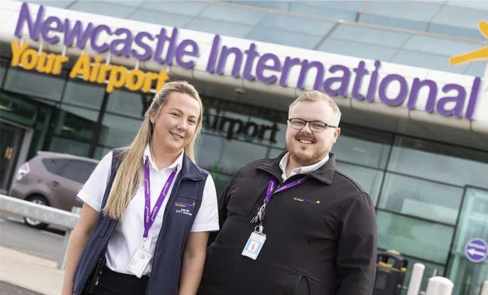 Newcastle College alumni Charlotte and Aaron smiling at the camera standing outside Newcastle International Airport.