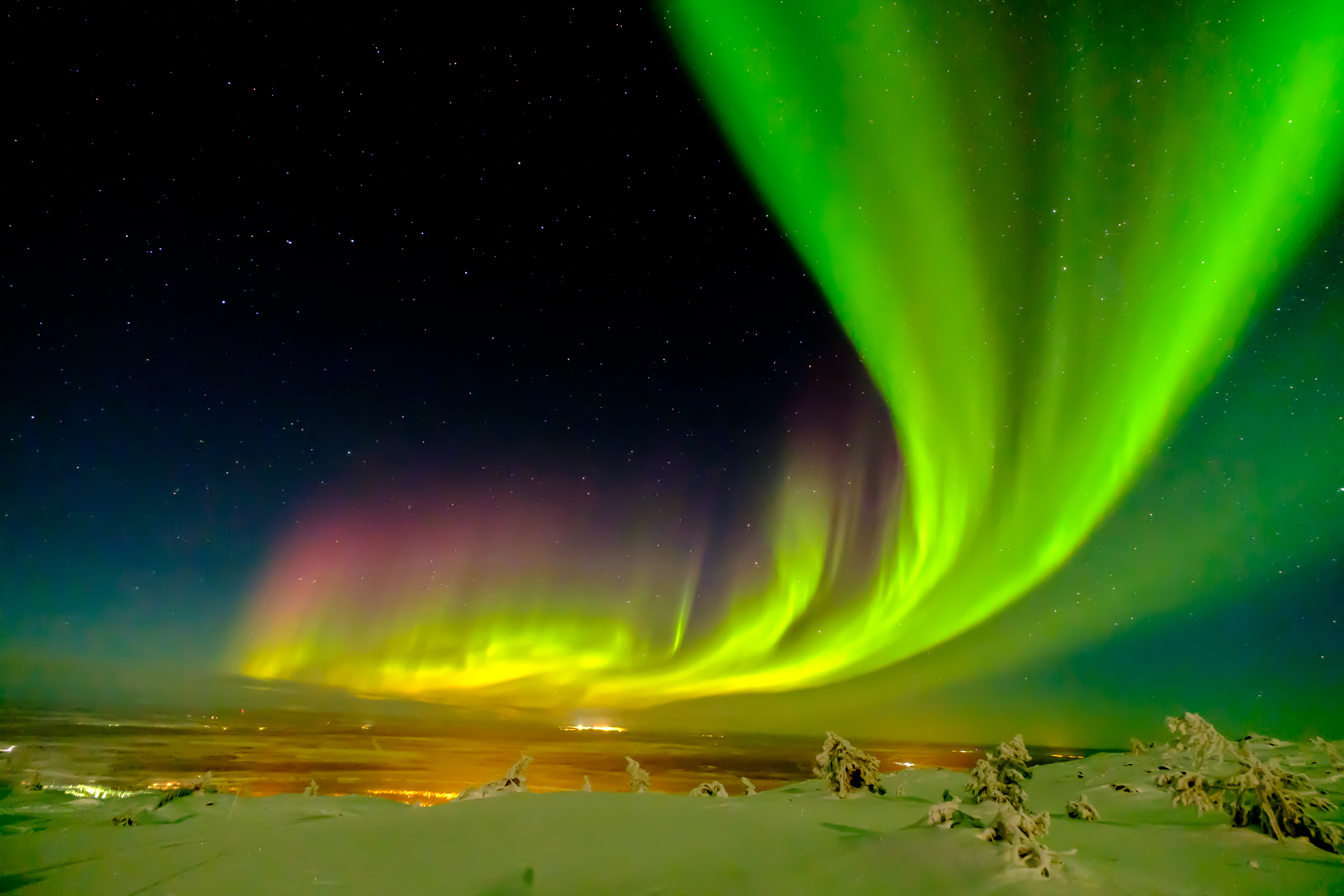 Northern lights in Lapland. The colours of the Northern lights are a blend of green, yellow, orange and pink. The Northern lights are above a snowy landscape with the snow covering the fields and trees. The photo was taken at night and you can visually see the stars in the sky.