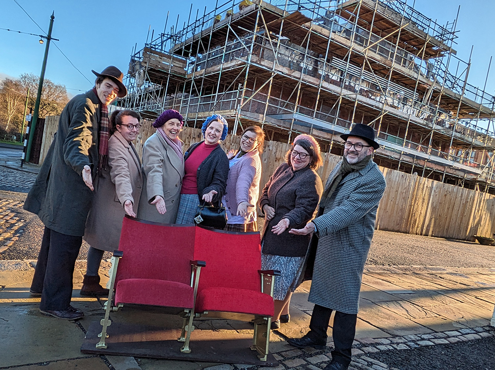 Group of people gathered around two traditional red cinema chairs and pointing at them in front of a scaffolded building.