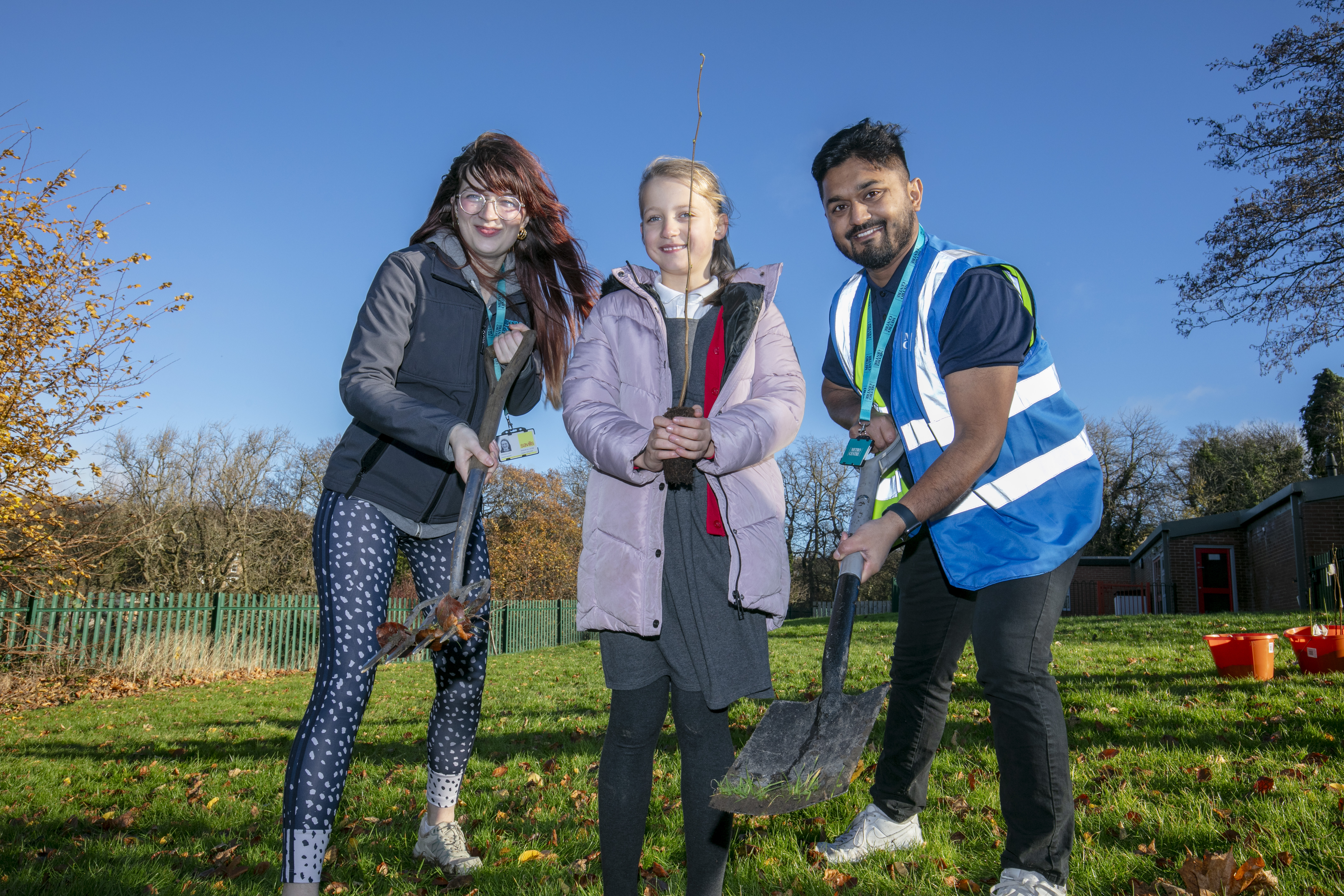 Three people stood in a grassy field . On the far left a woman is holding a long trowel with leaves attached, in the middle a young girl is holding a tree sprout. On the right a man is in a blue hi-vis holding a shovel.