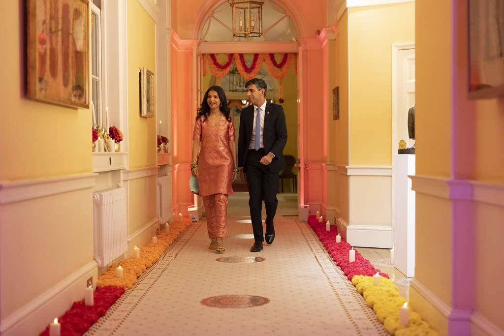 Rishi Sunak and his wife walking hand-in-hand down the No. 10 corridor looking at the paintings on the wall and smiling. The corridor is lined with flowers and candles.