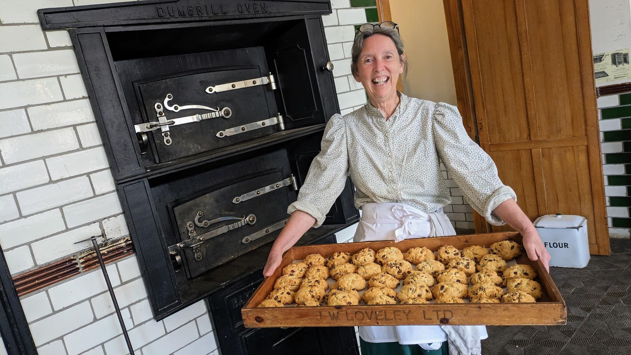 Woman in retro clothing holding a tray of baked scones in front of an arger.