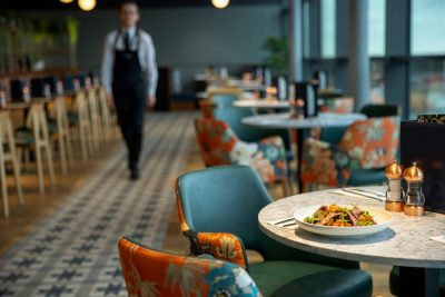 Image of food on one of the restaurant tables and waiter walking to the left of the image in between the tables.