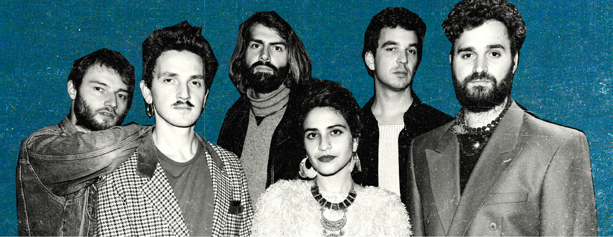 Group of six musicians stood in front of blue background looking towards the camera.